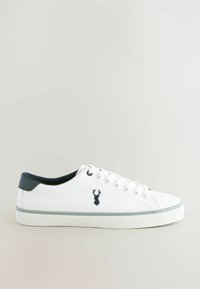 Witte canvas sneaker met donkerblauwe hielaccent, platte rubberen zool en geborduurd hertlogo aan de zijkant. Zes gaatjes voor veters.