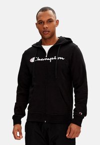 Felpa nera con zip realizzata in tessuto morbido, con cappuccio regolabile tramite un cordino, caratterizzata dal logo bianco "Champion" sul petto e tasche frontali.
