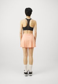 Zwarte sportbeha met netdetails, gecombineerd met een licht perzikkleurige skort. Persoon draagt witte sokken en zwarte sportschoenen. Transparante achtergrond.