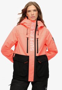 Jasny koralowy i czarny wodoodporny płaszcz z kapturem, z zamykanymi na zamek kieszeniami, dużym logo z przodu i kontrastującymi akcentami.