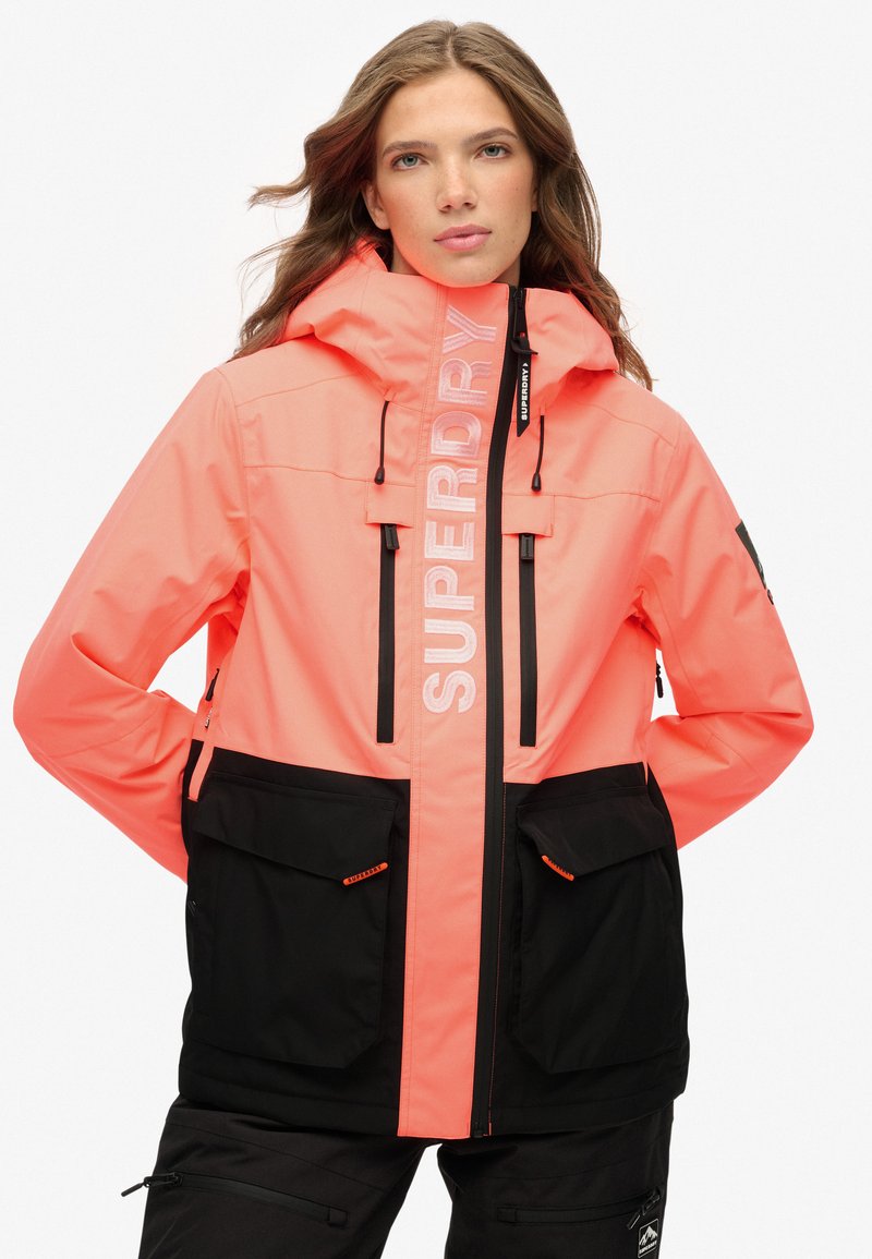 Jasny koralowy i czarny wodoodporny płaszcz z kapturem, z zamykanymi na zamek kieszeniami, dużym logo z przodu i kontrastującymi akcentami.