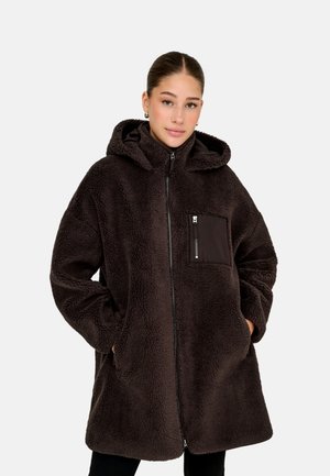 Jeune femme portant une veste à capuche oversize en polaire sherpa marron foncé avec fermeture éclair et poche poitrine, mains dans les poches latérales, fond neutre.