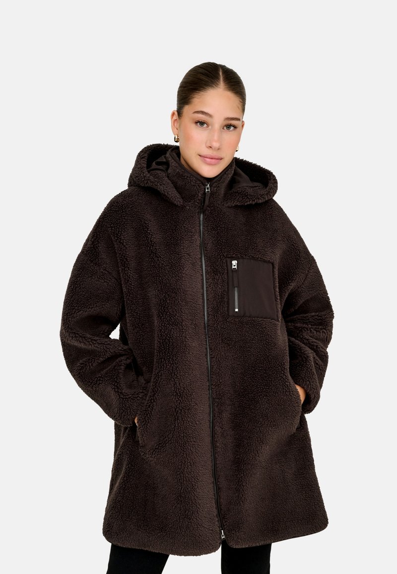 Jeune femme portant une veste à capuche oversize en polaire sherpa marron foncé avec fermeture éclair et poche poitrine, mains dans les poches latérales, fond neutre.