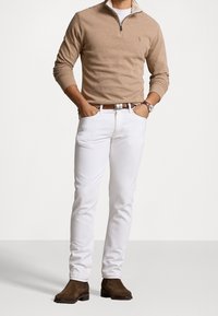 Světle béžový svetr na zip, bílé slim-fit džíny a hnědé semišové kotníkové boty. Texturovaný materiál a kontrastní barvy v celém outfitu.