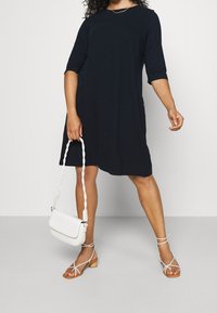 Marinblå T-shirtklänning med trekvartsärmar, sidofickor, mjuk textur, kombinerad med vita sandaler med remmar och en vit handväska.