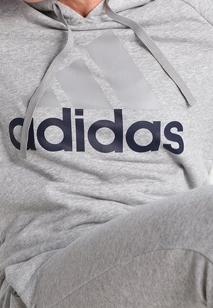 Osoba nosząca jasnoszarą bluzę z kapturem Adidas oraz dopasowane spodnie dresowe z widocznym logo na klatce piersiowej.