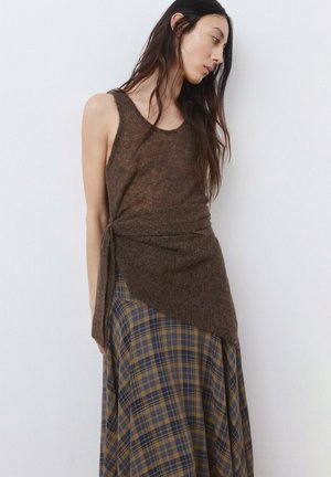 Mujer con cabello largo y oscuro, vestida con una blusa marrón sin mangas tipo envolvente y una falda de cuadros azules y marrones, de pie contra una pared blanca.