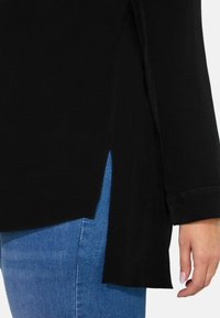 Czarna bluzka z długim rękawem i bocznymi rozcięciami, wykonana z teksturowanej tkaniny, z mankietami. Noszona na jasnoniebieskie dżinsy, ukazująca detale u dołu.