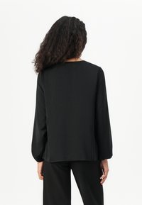 Vrouw met lang krullend haar, gekleed in een zwarte blouse met lange mouwen en zwarte broek, staand met haar rug naar de camera tegen een witte achtergrond.