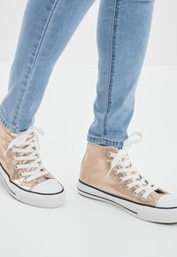 High-top sneakers made of metallic gold material, featuring white laces and rubber soles with black accents, paired with light blue jeans.