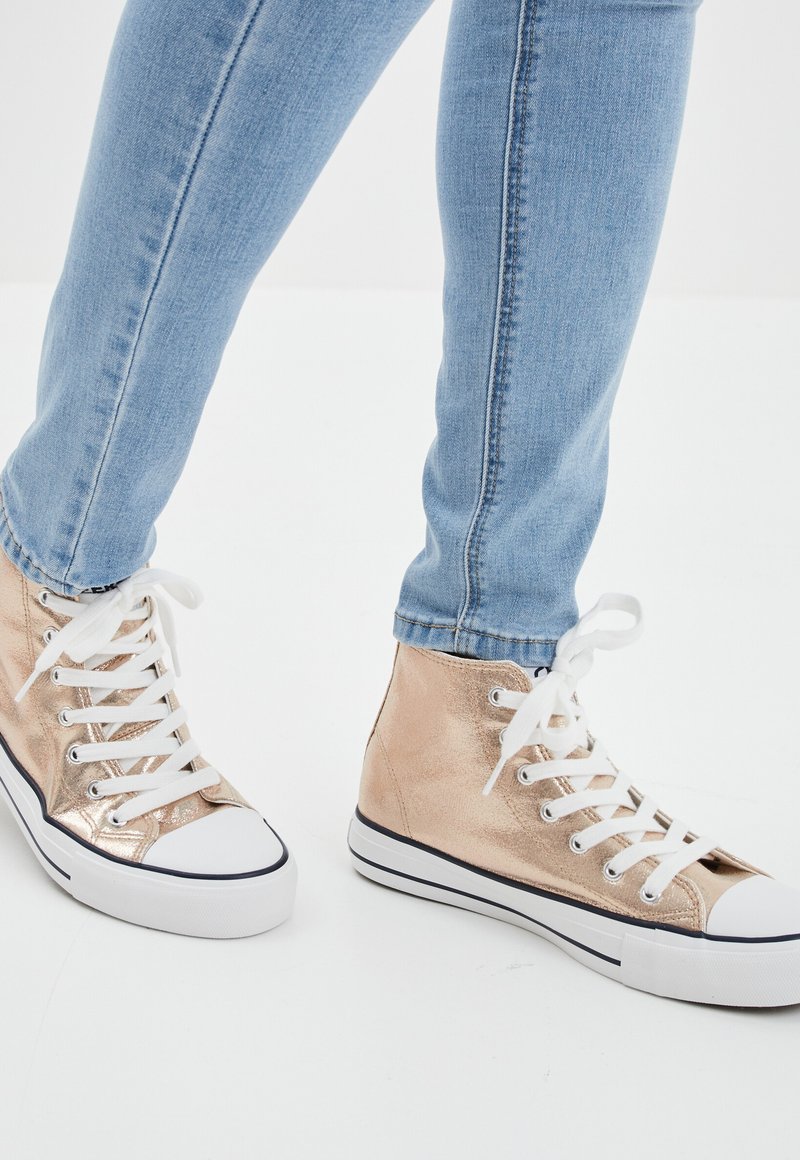 High-top sneakers made of metallic gold material, featuring white laces and rubber soles with black accents, paired with light blue jeans.