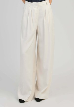 Pantalon blanc à jambes larges avec plis sur le devant et taille, porté avec des chaussures noires pointues et un haut sombre rentré.