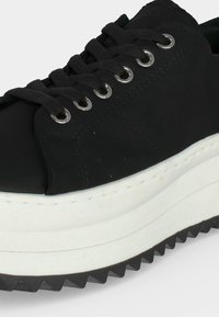 Czarny sneaker na sznurówki z grubą białą podeszwą platformową i żłobioną czarną podeszwą zewnętrzną, zbliżenie na bok i przednią część buta.