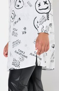 Chemise blanche avec des graphismes noirs présentant des émoticônes et du texte, associée à un pantalon en cuir noir. La chemise a une coupe décontractée et des fentes sur les côtés.