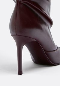 Burgundské vysoké podpätky s elegantným koženým povrchom, špicatou špičkou a zúženým stiletto podpätkom. Obsahuje jemné drapérie z kože.