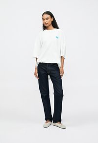 Tricou alb supradimensionat cu grafică albastră, blugi din denim închis la culoare și pantofi slip-on deschiși la culoare, pe un fundal simplu.