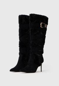 Stivali neri in suede al ginocchio con fodera in pelliccia sintetica, punte affusolate, tacchi a stiletto e cinturini decorativi avvolgenti con fibbia dorata.