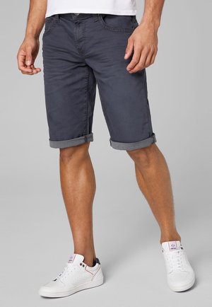 Mężczyzna w ciemnych podwiniętych szortach i białych trampkach, stojący na jednolitym szarym tle.