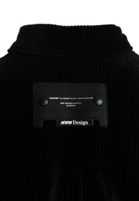 Tela de pana negra con un alto cuello. Presenta una etiqueta rectangular de cuero con texto en relieve y dos botones negros.