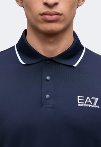 Άνδρας που φοράει μπλουζάκι πόλο Emporio Armani EA7 σε ναυτικό μπλε χρώμα με λευκή επένδυση στο γιακά και τρία μαύρα κουμπιά.
