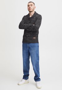 Dunkelgrauer gestrickter Pullover mit Schalkragen, ausgestattet mit einem Knopfverschluss und einem kleinen Lederetikett. Kombiniert mit hellblauen, geraden Jeans.