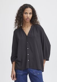 Black blouse with a V-neck and button front, featuring loose, cropped sleeves and a slightly textured fabric. Worn with blue denim trousers.