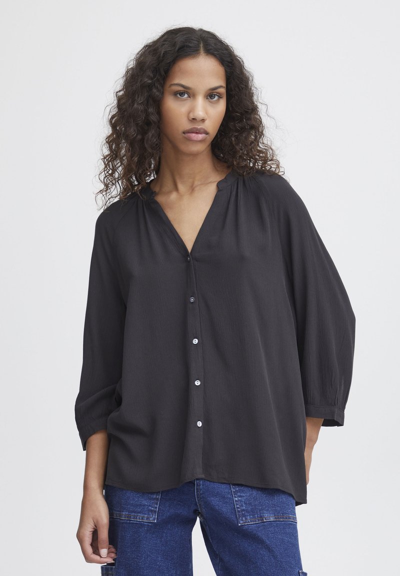 Black blouse with a V-neck and button front, featuring loose, cropped sleeves and a slightly textured fabric. Worn with blue denim trousers.