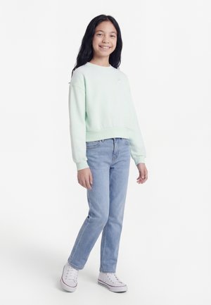 Junges Mädchen lächelt, trägt einen hellgrünen Pullover, blaue Jeans und weiße Turnschuhe, steht vor einem weißen Hintergrund.