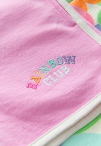 Ružová látka má vyšívaný nápis "RAINBOW CLUB" v mnohých farbách. Okraje sú zakončené bielym lemom, textúra vyzerá mäkká a hladká.