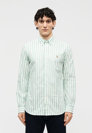 Νεαρός άνδρας με κοντά σγουρά μαλλιά, φορώντας ένα λευκό πουκάμισο με πράσινες κάθετες ρίγες και σκούρο παντελόνι, στέκεται μπροστά σε ένα απλό φόντο.