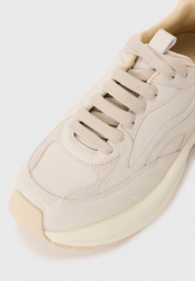 Beige sneaker with a smooth leather upper, textured accents, flat laces, and a cushioned sole with a slight curve at the toe.