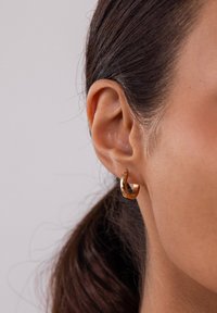 Goldene Creole mit polierter Oberfläche, die ein dünnes Design und eine subtile Kurve aufweist, getragen am Ohrläppchen einer Person.