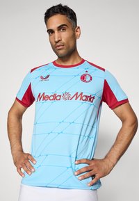 Vīrietis, kas ģērbies gaiši zilā un sarkanā Feyenoord Rotterdam futbola kreklā ar "Media Markt" logotipu, stāv ar rokām uz gurniem uz viendabīga fona.