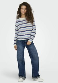 Stickad tröja med blå och krämfärgad vågmönster, rund halsringning och lång ärm, i kombination med vida jeans och vita sneakers.
