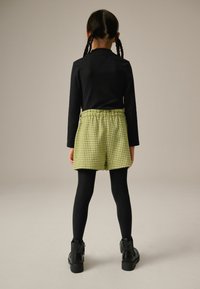 Sort ribbet langærmet top, gule ternede shorts med elastisk talje, sorte leggings og sorte ankelstøvler. Ensfarvet baggrund.