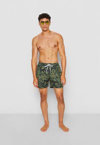 Männer Badehosen mit einem grünen Paisley-Muster, elastischem Bund, weißem Kordelzug und leichtem Stoff. Getragen mit einer grünen Sonnenbrille.