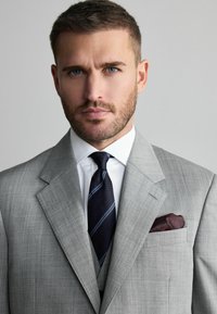Traje gris claro con un diseño estructurado, camisa blanca y una corbata azul marino con detalles a rayas. Un pañuelo burdeos añade un toque de detalle.