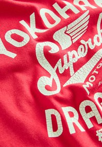 Rød bomulds T-shirt med revnet hvid tekst og grafik, herunder vinger og brandnavn, med en blød, glat tekstur.