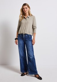 Beige knäppt blus med långa ärmar och dragsko i nederkant, i kombination med vida blå denimjeans. Modell i svarta platta skor.