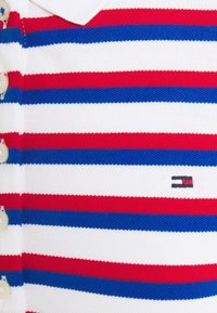 Gros plan d’un tissu blanc avec des rayures horizontales rouges et bleues et des boutons crème sur le côté gauche, avec un petit logo rectangulaire.