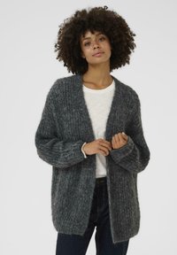 Siv cardigan iz debele pletenine z odprtim sprednjim delom, širokimi rokavi in teksturno vzorcem. Usklajen z belim srajcem in temnimi jeans hlačami.