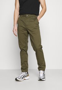 Olijfgroene broek met een rechtliggende snit, gemaakt van katoen, met een enkele knoopsluiting en zijzakken. Draagt bij zilveren sneakers.