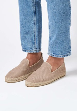 Des chaussures espadrilles beiges avec une texture tissée, un côté élastique et une semelle en jute, portées avec un jean en denim léger bleu retroussé.