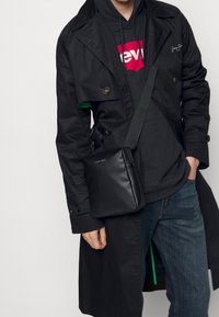 Trench coat nero doppiopetto, felpa nera con logo rosso, jeans blu e una piccola borsa a tracolla in pelle nera. Dettaglio interno verde visibile.