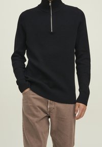 Pull en maille côtelée noire avec un col montant et une fermeture éclair à moitié. Le matériau semble doux avec des détails texturés le long du corps.