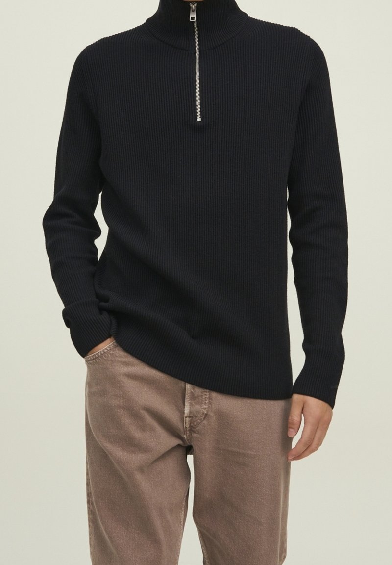 Pull en maille côtelée noire avec un col montant et une fermeture éclair à moitié. Le matériau semble doux avec des détails texturés le long du corps.