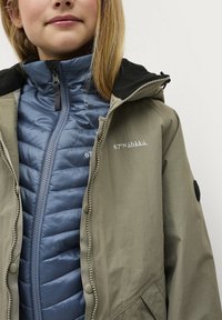 Lichte olijfgroene jas met een capuchon, gecombineerd met een blauwe gewatteerde binnensjas met een rits aan de voorkant en logo-details.