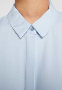Gros plan d'une personne portant une chemise à col texturée bleu clair, boutonnée au niveau du col.