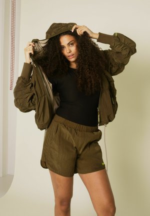 Femeie ajustând gluga unei jachete largi verde măsliniu, purtând pantaloni scurți asortați și un top negru, stând în fața unui fundal simplu și deschis la culoare.
