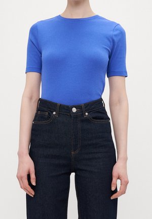 Person trägt ein eng anliegendes blaues Kurzarmshirt, das in hochtaillierte dunkelblaue Jeans mit sichtbaren Nähten und Fronttaschen gesteckt ist.